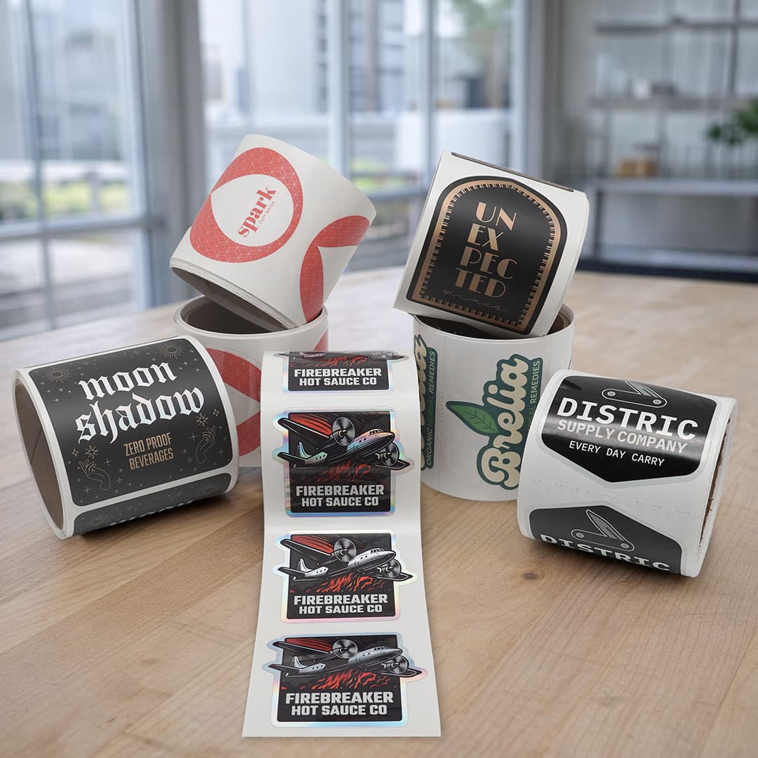 Assortment of custom kiss cut roll stickers featuring multiple brand designs displayed on a tabletop in a modern workspace.