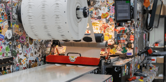 A large roll of label material feeds through a sticker-covered production machine in a print shop.