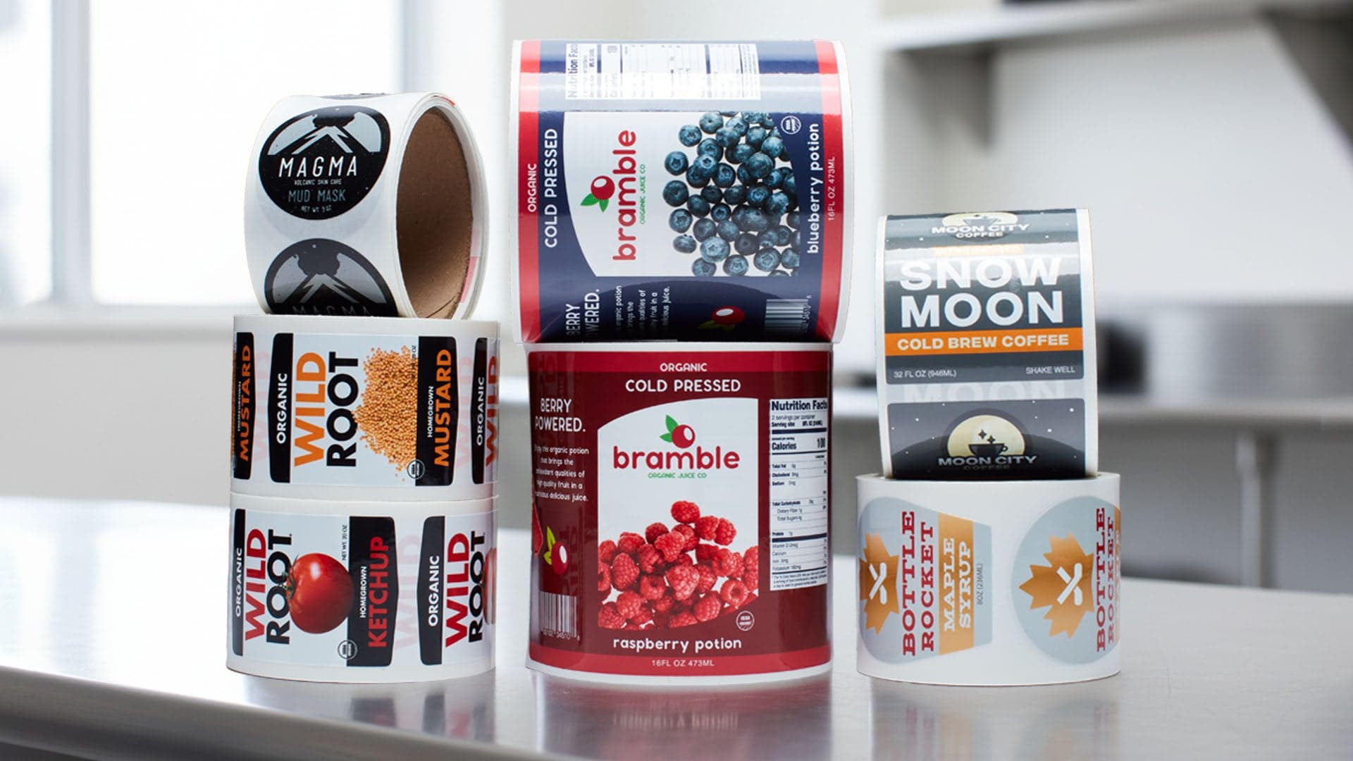 Rolls of custom product labels for food and beverage brands are stacked on a counter, including coffee, ketchup, mustard, and cold-pressed juice designs.
