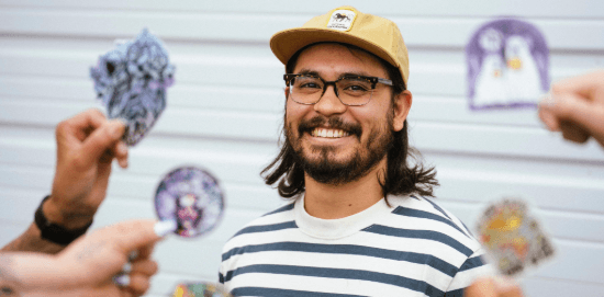 A smiling person in glasses and a yellow hat stands while several hands hold colorful stickers in the foreground.