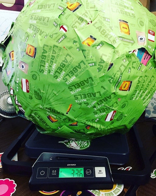Large green sticker-covered ball sitting on a digital scale, showing a weight of 7.36 pounds, with overlapping product label stickers visible on the surface.