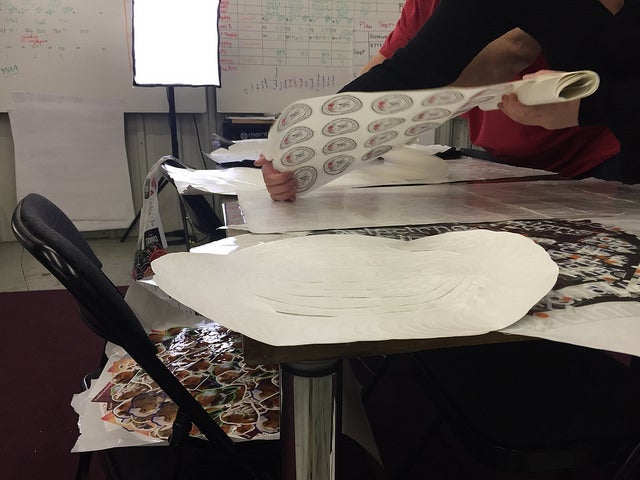People working at a table covered with large sticker sheets, lifting and arranging full pages of printed stickers under bright studio lighting, with additional sticker sheets spread across the workspace.