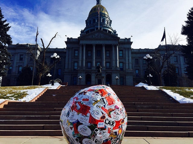 arge sticker-covered ball displayed on the steps leading up to the Colorado State Capitol building, with the dome and front facade visible in the background under a blue sky.