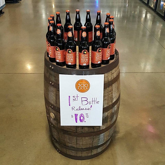 Display of Slow Order Stout bottles arranged on a wooden barrel with a sign announcing 300 Suns Brewing’s first bottle release.