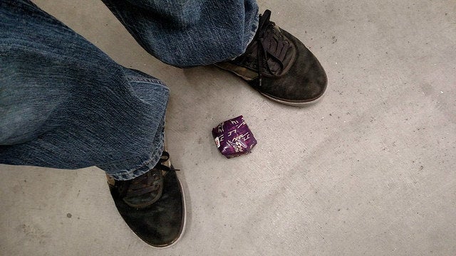 Person wearing dark shoes and blue jeans standing on a concrete floor with a small crumpled purple object between their feet.