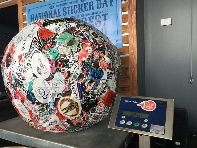 Large sticker-covered ball displayed on a metal scale during an official weigh-in, with the digital screen showing 231.6 pounds and a National Sticker Day banner in the background.