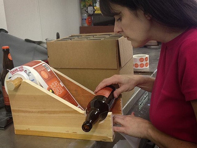 Person applying a custom label by hand to a beer bottle using a wooden labeling jig, with rolls of StickerGiant-printed labels nearby.