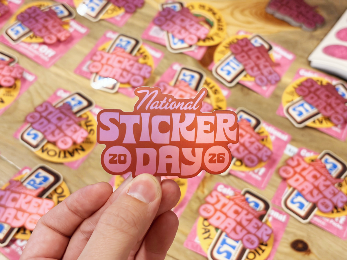 A hand holds a pink &lsquo;National Sticker Day 2026&rsquo; sticker above a table covered with similar stickers.