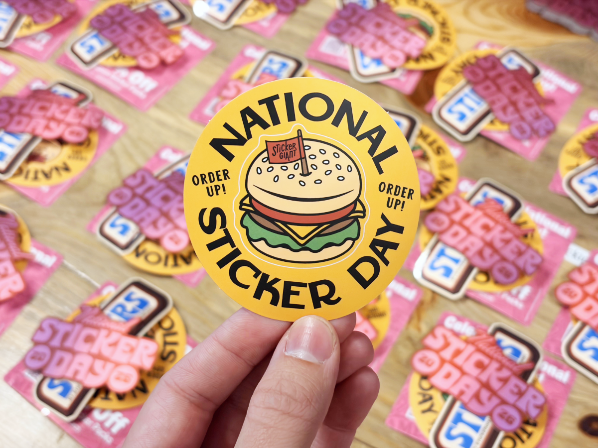 A hand holds a round yellow &lsquo;National Sticker Day&rsquo; burger-themed sticker above a table covered with similar sticker designs.