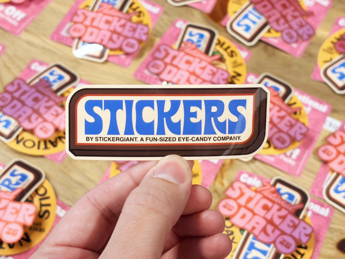 A hand holds a retro-style &lsquo;Stickers by Stickergiant&rsquo; sticker above a table covered with National Sticker Day stickers.