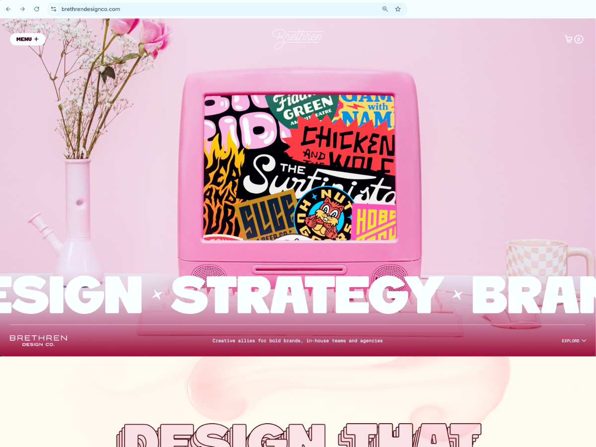 A pink retro computer displaying colorful graphic logos sits on a pink desk between a flower vase and a checkered mug on a pastel pink background.