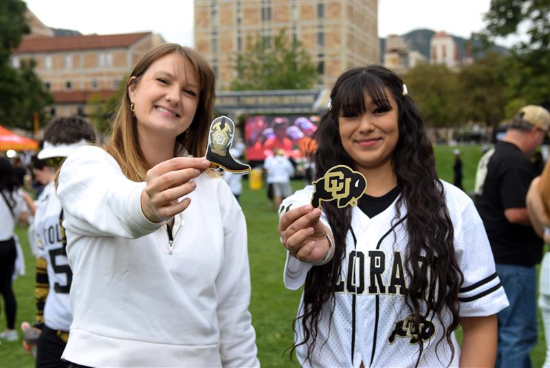 Two smiling women hold up University of Colorado stickers at an outdoor event on campus.