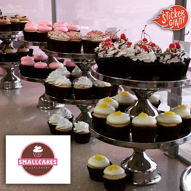 Tiered metal stands filled with assorted frosted cupcakes inside a Smallcakes: A Cupcakery bakery display, with the Smallcakes logo and a StickerGiant logo shown in the image.