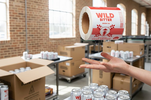 Person tossing a roll of &lsquo;Wild Bitter&rsquo; beer labels above a table of canned beverages in a bright brewery packing area.