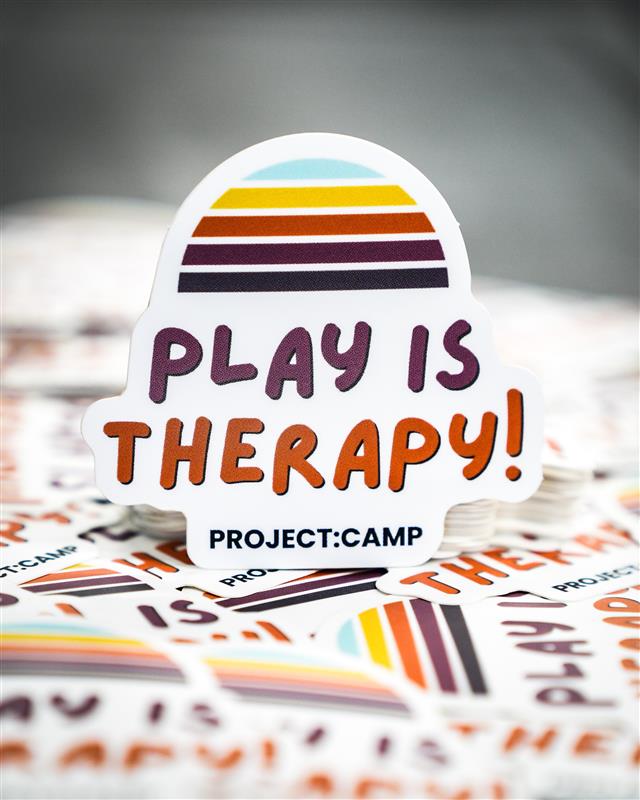 A die-cut sticker reading &ldquo;PLAY IS THERAPY!&rdquo; with retro stripes above and &ldquo;PROJECT:CAMP&rdquo; below.