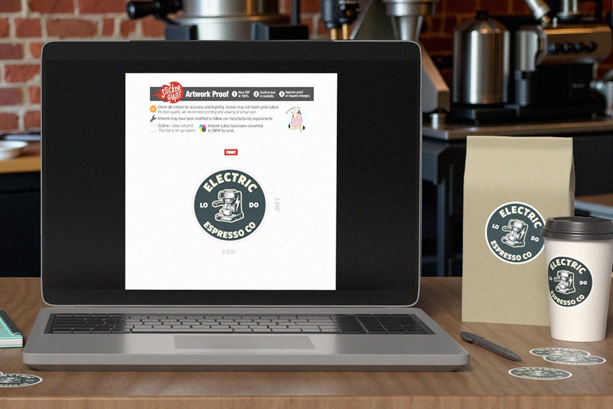 Laptop displaying a StickerGiant artwork proof for a round coffee logo, shown on a caf&eacute; counter with matching labeled coffee bag, cup, and stickers.