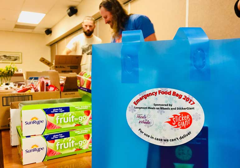 Blue emergency food bag labeled “Emergency Food Bag 2017,” with StickerGiant and Meals on Wheels logos, shown on a table while volunteers pack food items in the background.