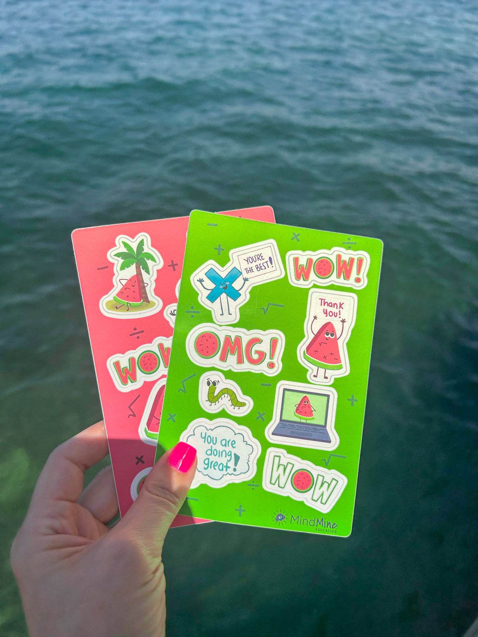 A hand holds two colorful sticker sheets with playful characters and phrases like &ldquo;OMG,&rdquo; &ldquo;WOW,&rdquo; and &ldquo;You are doing great!&rdquo; against a backdrop of blue water.