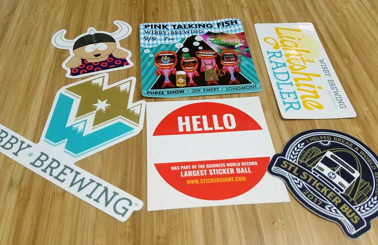 Assorted custom stickers from Wibby Brewing and StickerGiant displayed on a wood surface, including event stickers, logo stickers, a name-tag style sticker, and a Guinness World Record StickerGiant design.