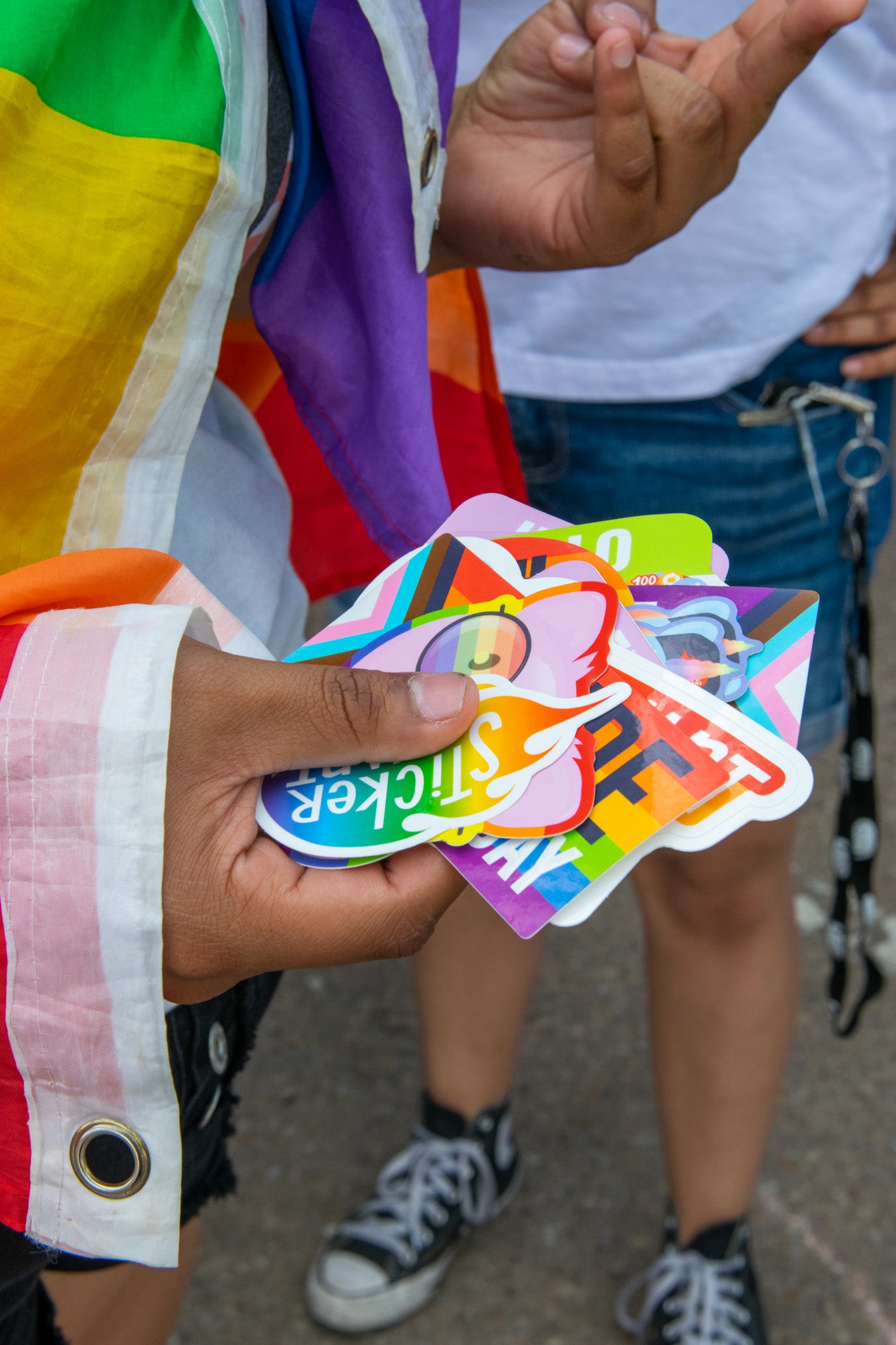 A person draped in a rainbow flag holds a handful of colorful Pride-themed stickers while standing with others at an outdoor event.
