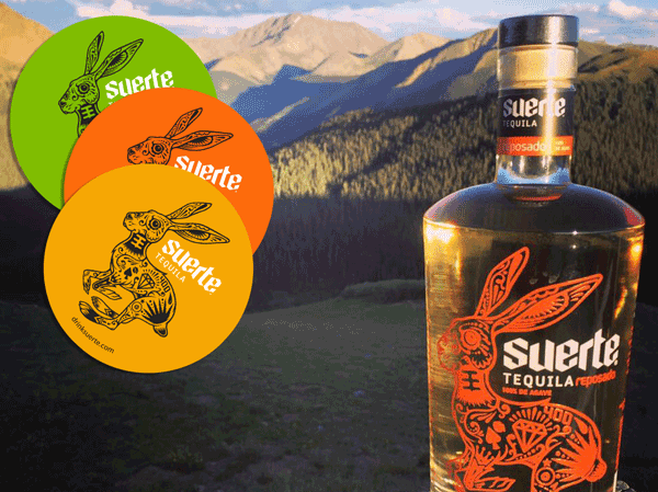 Suerte Tequila bottle with branded rabbit stickers in front of a mountain landscape.