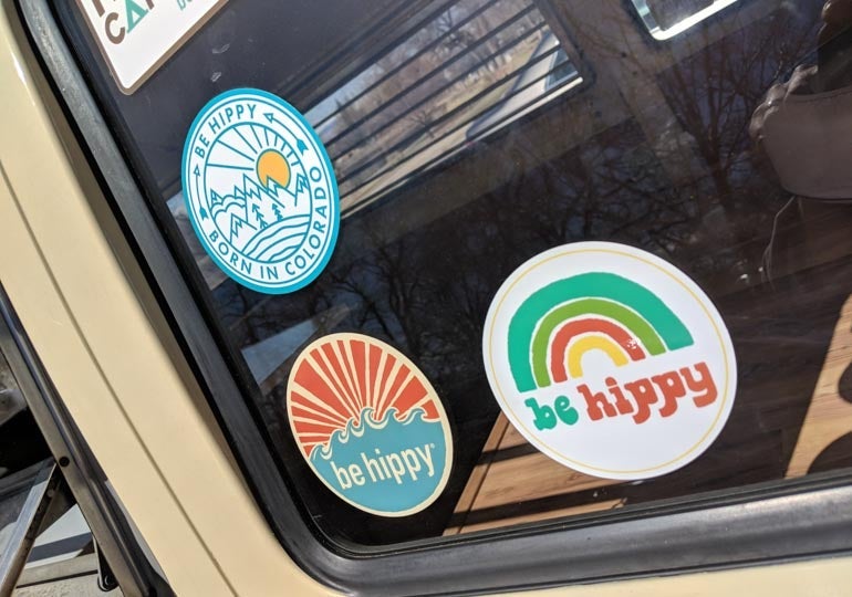 Three Be Hippy stickers displayed on the window of a vehicle, featuring a Colorado mountain design, a retro wave design, and a rainbow graphic, all printed as custom Die Cut Stickers