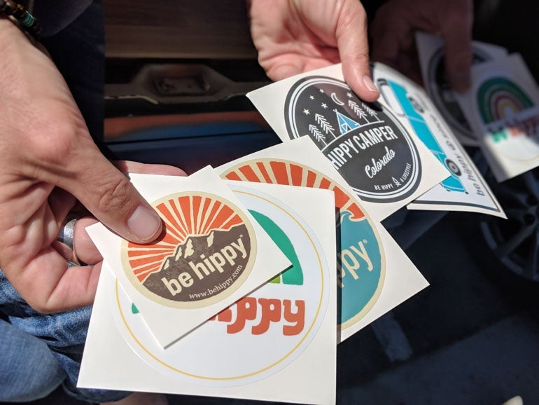Two hands holding a variety of Be Hippy stickers, including retro mountain designs, rainbow graphics, and a “Hippy Camper” illustration, all printed as custom Die Cut Stickers.