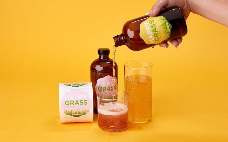 Hand pouring branded “Happy Grass” beverage into glasses with labeled bottles nearby.