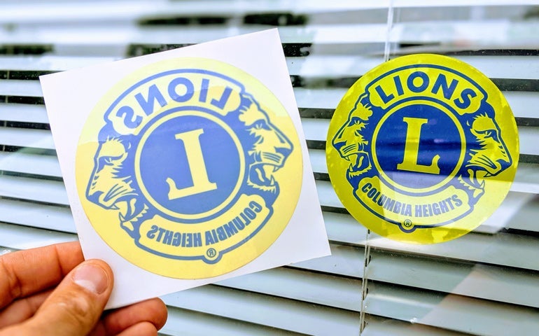 Clear Lions Club sticker shown on backing and applied to a window.