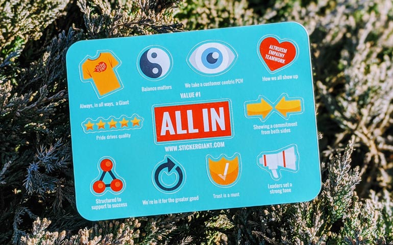 Sticker sheet featuring StickerGiant’s “All In” Core Value with icons representing balance, teamwork, trust, pride, leadership, and customer focus, displayed outdoors on greenery.