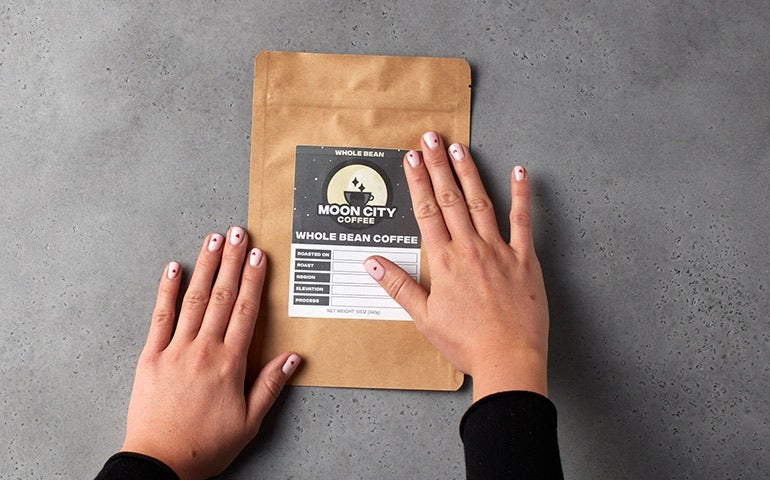 Hands applying a Moon City Coffee label to a kraft whole bean coffee bag.