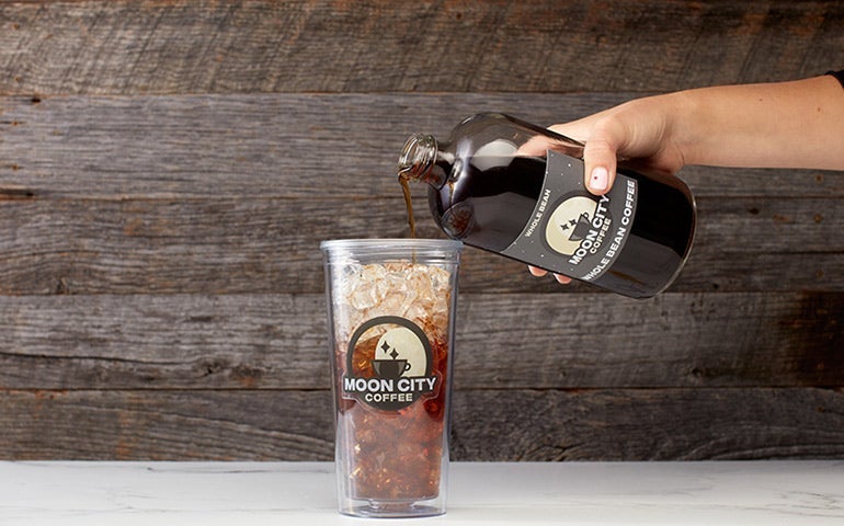 Hand pouring cold brew from a Moon City Coffee bottle into a branded iced cup.