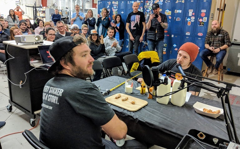 StickerGiant team members recreate a Hot Ones challenge as coworkers watch in a crowded warehouse setting.