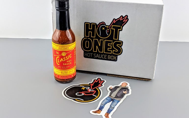 Hot Ones hot sauce bottle, branded subscription box, and two custom stickers featuring the Hot Ones logo and a host illustration displayed together on a gray surface.