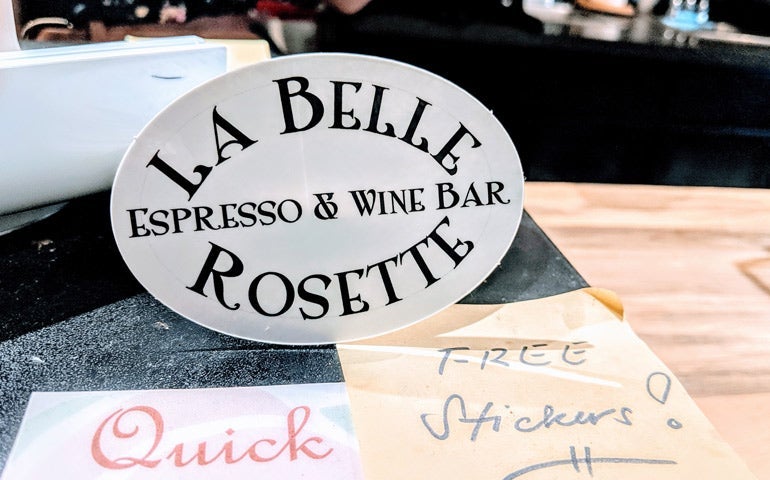 Oval sticker reading “La Belle Rosette Espresso & Wine Bar” displayed on a counter, with a handwritten note beside it offering free stickers.