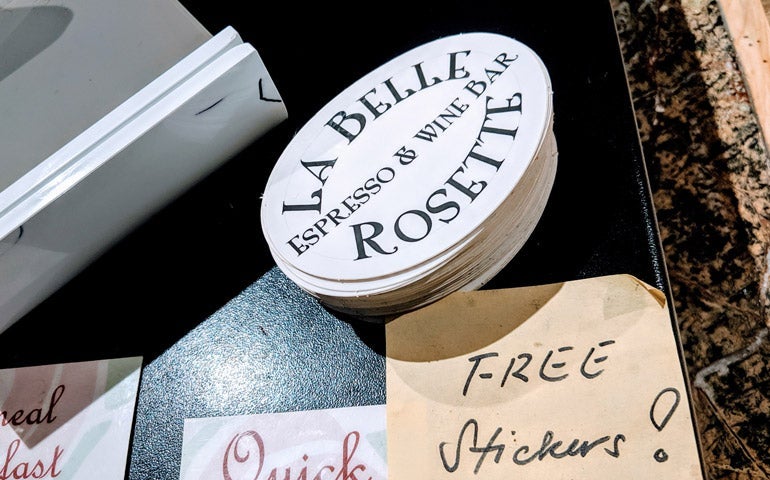 Stack of oval “La Belle Rosette Espresso & Wine Bar” stickers on a countertop beside a handwritten sign that reads “Free Stickers.”