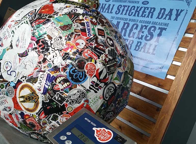 Saul, the World’s Largest Sticker Ball, covered in hundreds of colorful custom stickers and displayed on a scale during a National Sticker Day event, with a blue event poster in the background.