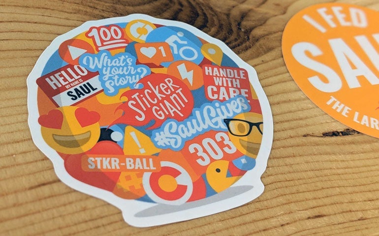 Colorful die cut sticker with Saul-themed icons and StickerGiant branding on a wooden surface.
