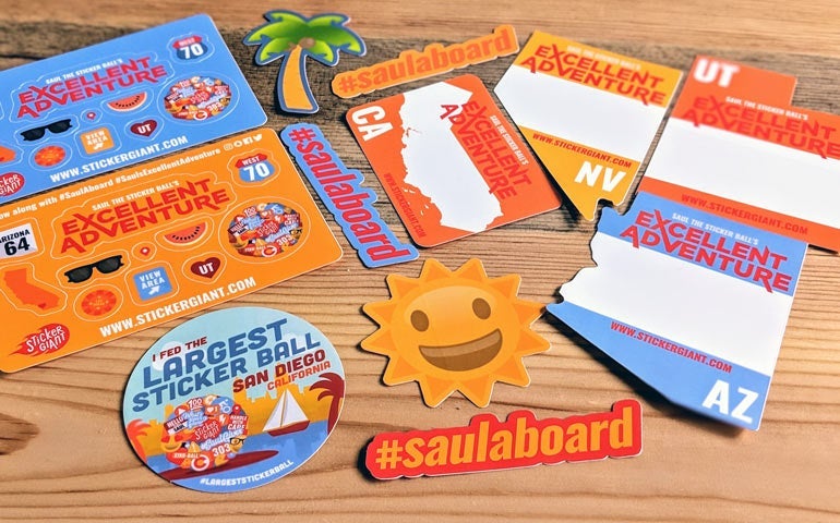 Assorted custom Excellent Adventure stickers, state-shaped stickers, hashtag decals, and a round Saul sticker arranged on a wooden surface.