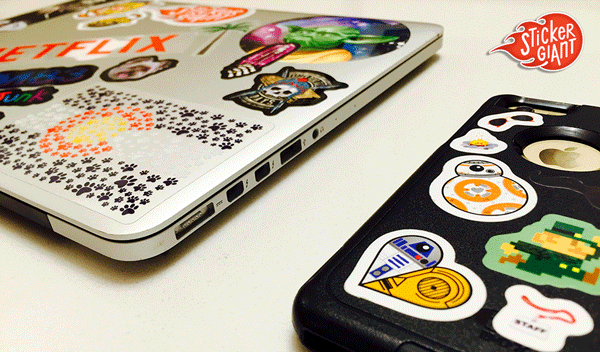 A laptop and phone covered in colorful custom stickers, including character designs and brand stickers, demonstrating how fans display stickers on personal devices.