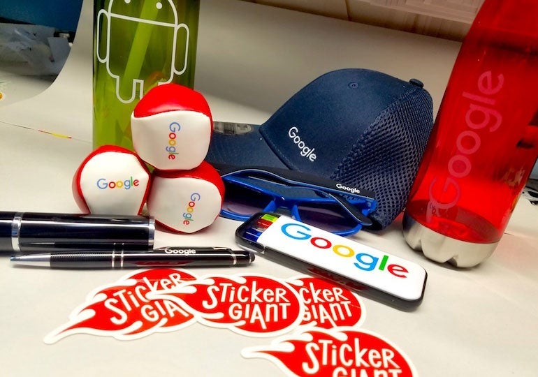 Assorted Google-branded swag items displayed on a table alongside several StickerGiant logo stickers.